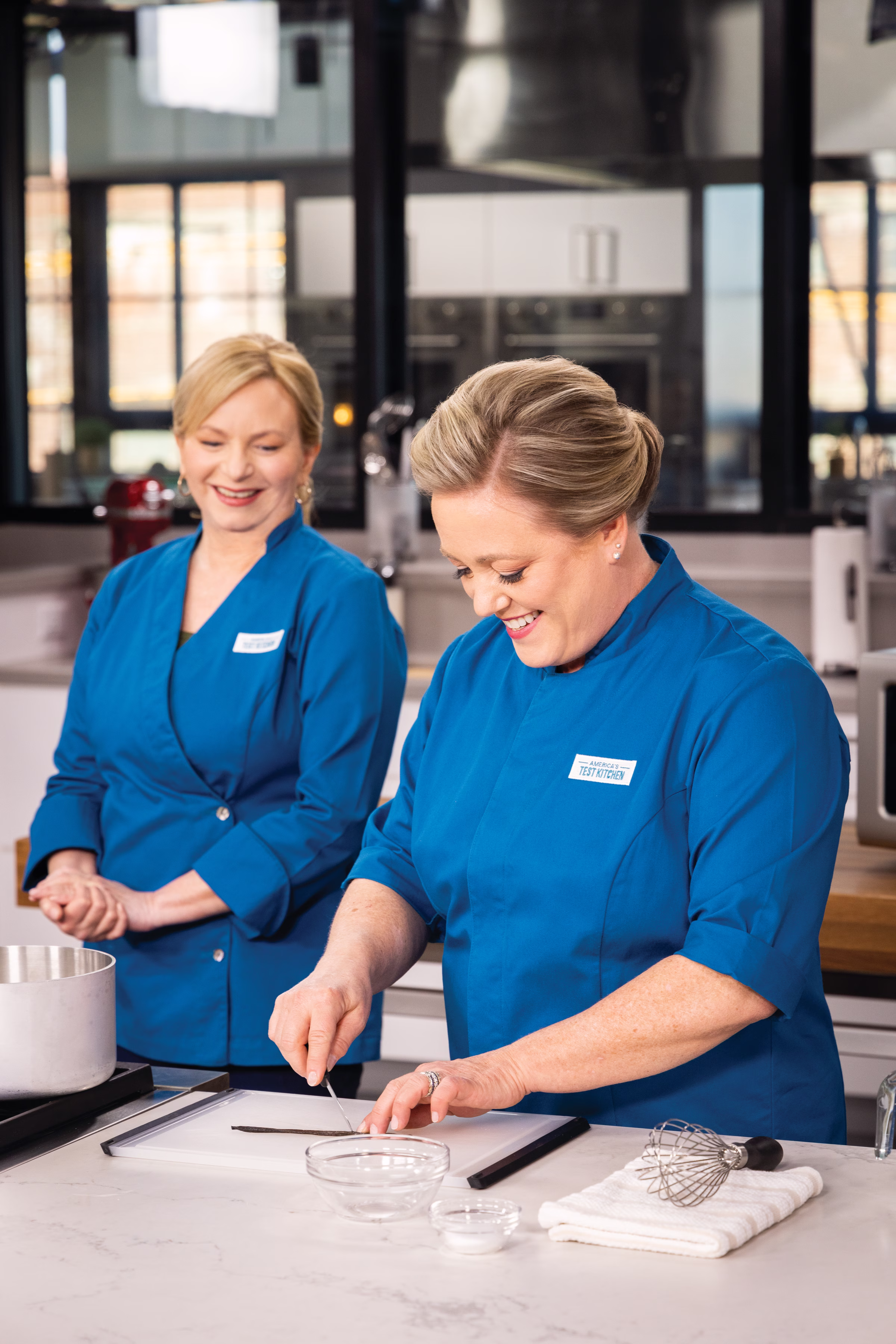 America's Test Kitchen hosts Bridget Lancaster and Julia Collin Davison preparing recipes in blue chef coats at the Boston test kitchen
