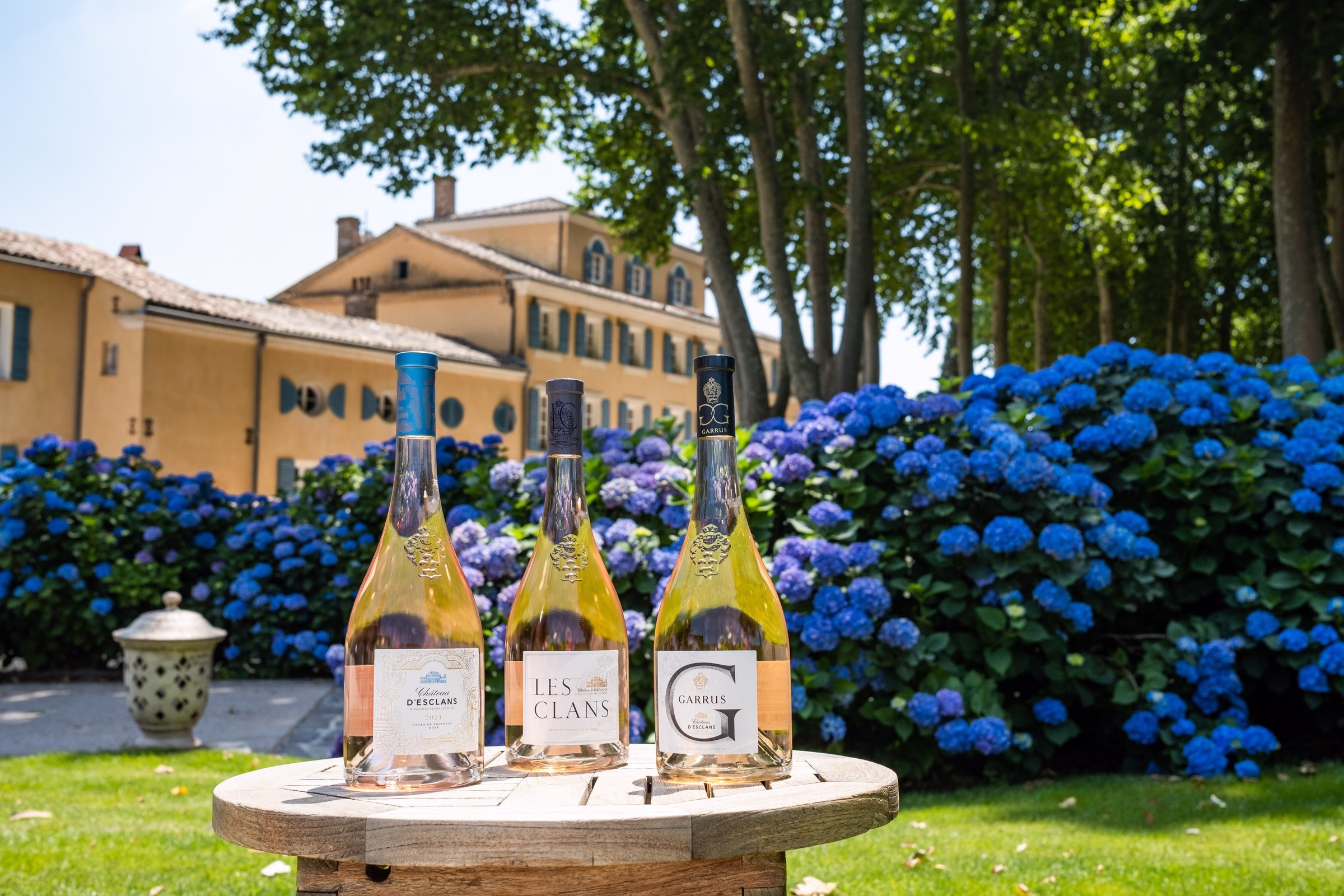 Chateau d'Esclans Les Clans and Garrus rosé bottle lineup on a barrel head with blue hydrangeas and chateau backdrop