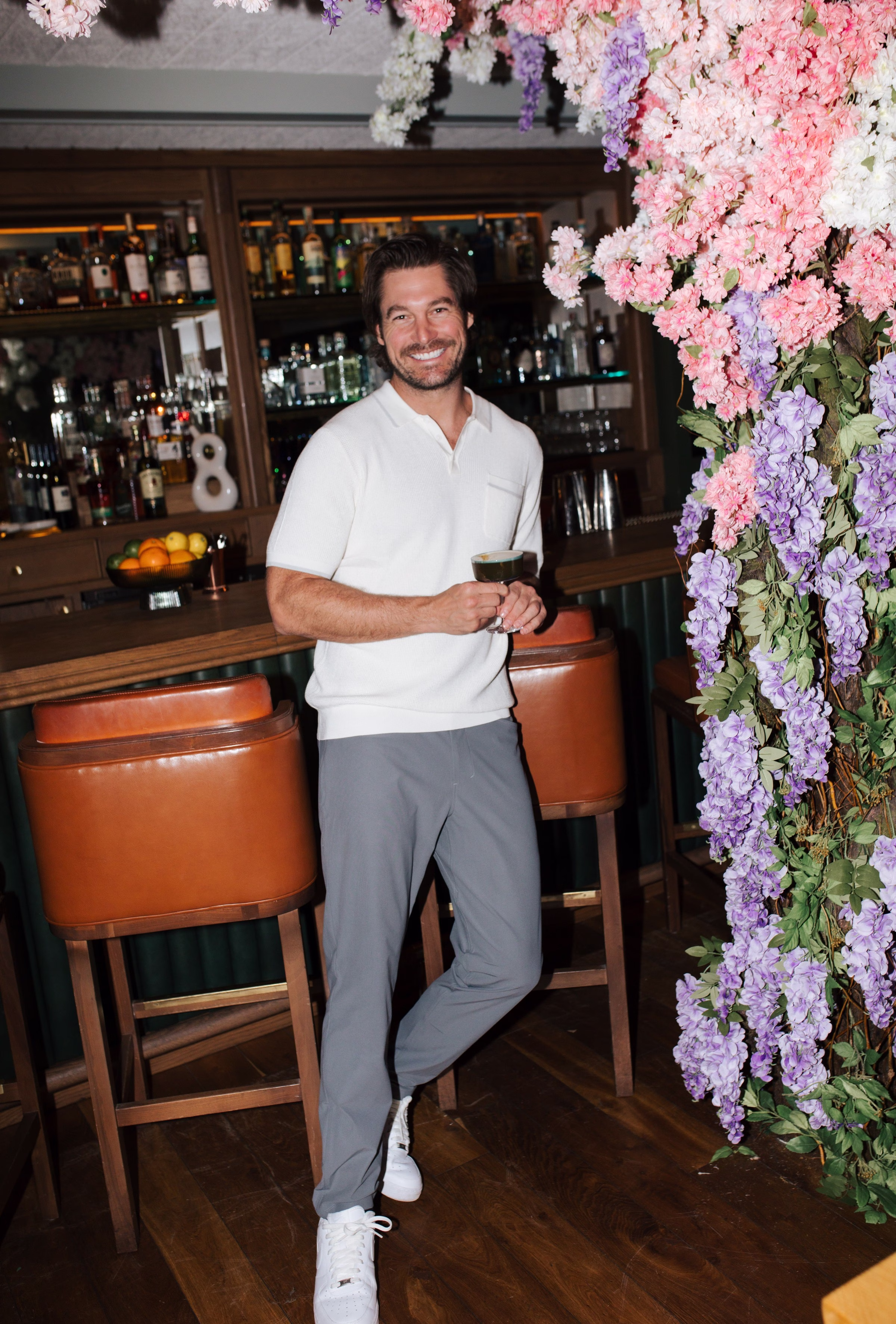 Craig Conover holding a cocktail glass, leaning against a bar counter surrounded by pink and purple floral installations at Hotel Emeline
