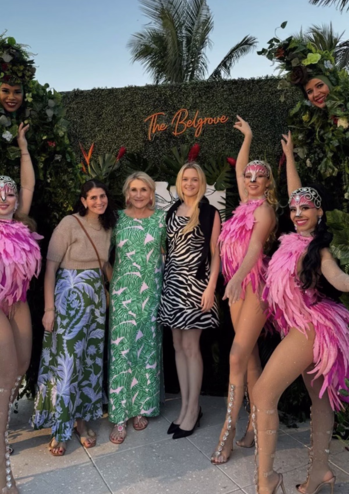 Group photo at The Belgrove venue featuring guests in tropical attire with costumed performers in pink feathers and face paint against hedge backdrop with palm trees