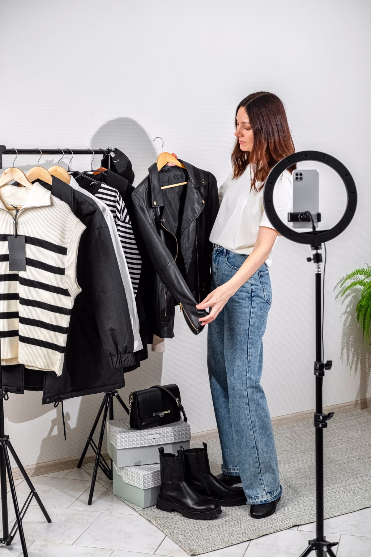 Female content creator selecting black leather jacket from clothing rack while filming fashion styling video with ring light and smartphone setup