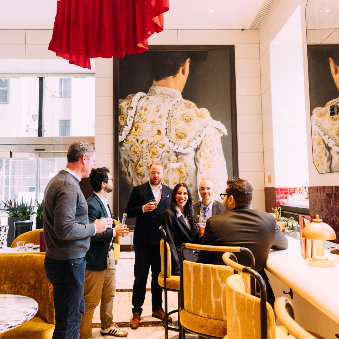 Group of professionals socializing with cocktails at Beast and Butterflies Spanish-inspired bar inside M Social Hotel New York Downtown Financial District, with large bullfighter mural and red cape installation overhead