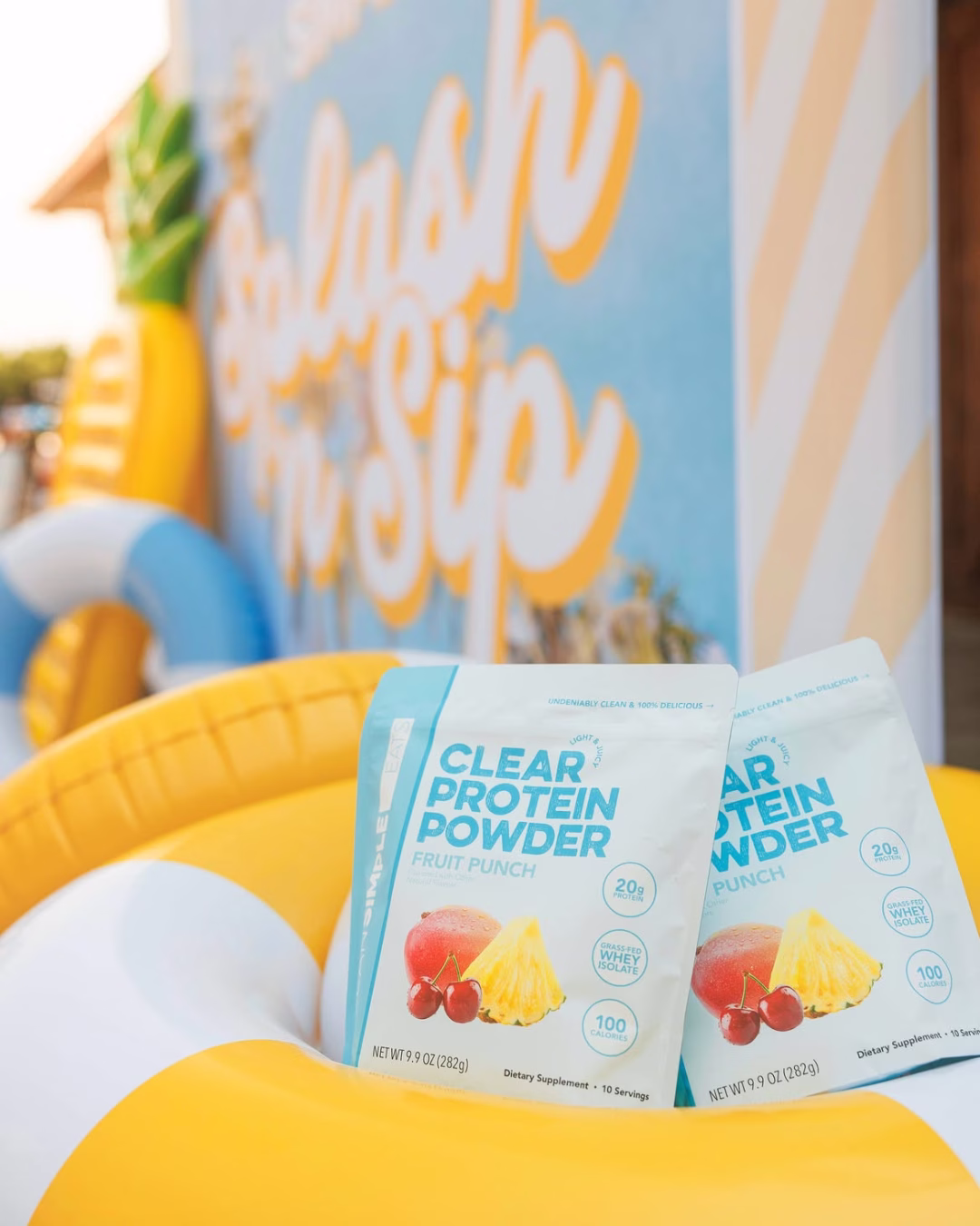 Two bags of Clean Simple Eats Clear Protein Powder in Fruit Punch flavor, featuring grass-fed whey isolate with 20g protein and 100 calories per serving, displayed on a yellow inflatable pool float at a summer Splash and Sip event