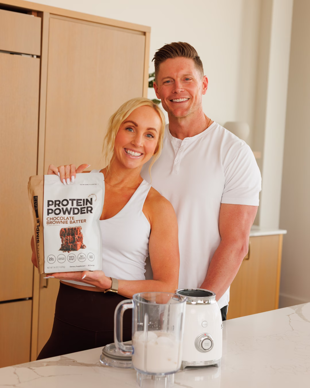 Clean Simple Eats founders Erika and JJ smile together in a modern kitchen while holding a bag of Chocolate Brownie Batter Protein Powder alongside a blender on a marble countertop