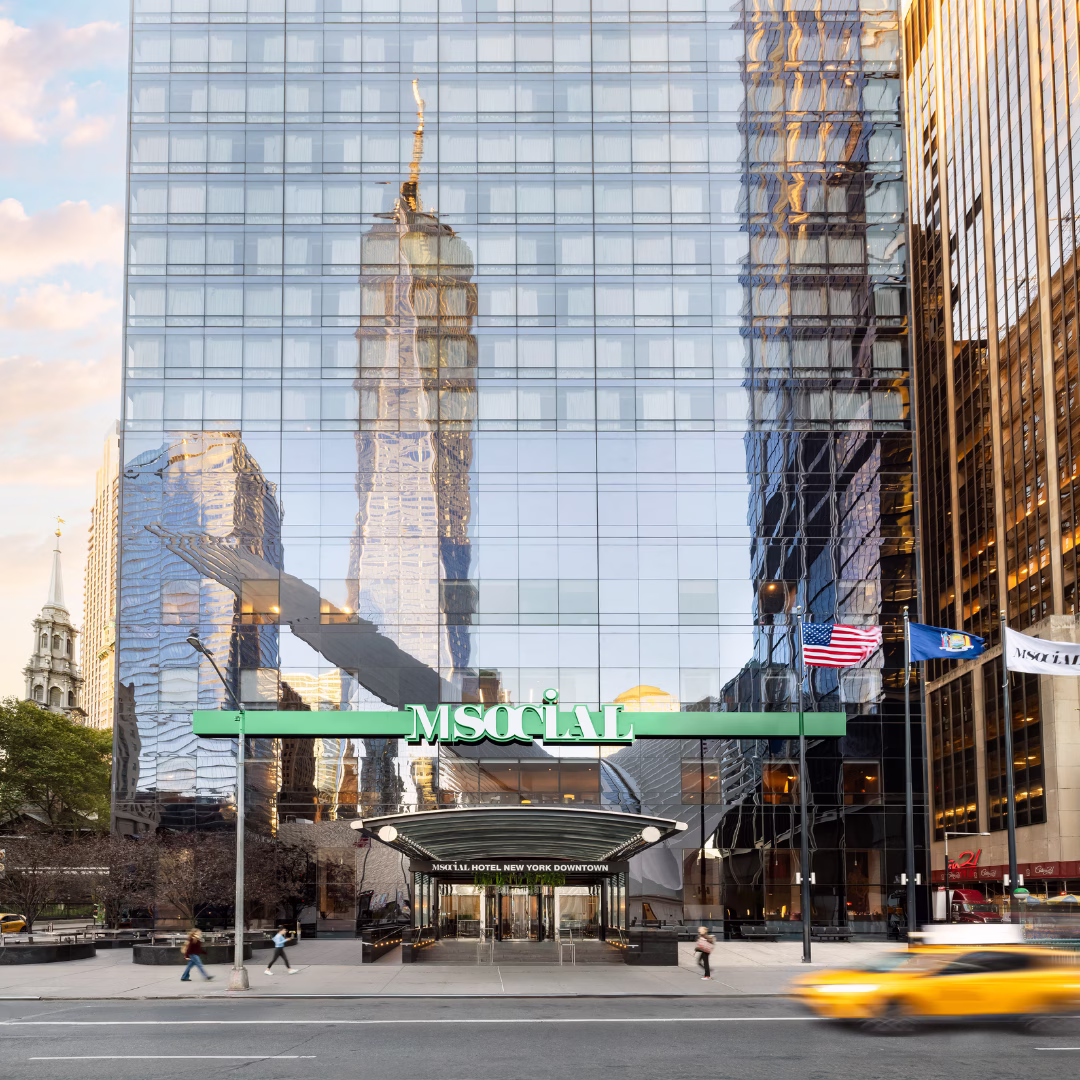 Exterior facade of M Social Hotel New York Downtown in the Financial District, featuring a glass curtain wall reflecting One World Trade Center, with a green branded canopy entrance, American and New York state flags, and a yellow taxi cab passing in the foreground