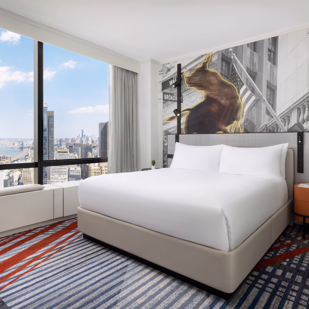 Bright king guest room at M Social Hotel New York Downtown featuring a bold NYC Financial District wall mural with the Charging Bull and Wall Street signage, striped red and blue carpet, and panoramic floor-to-ceiling windows overlooking the Manhattan skyline, Hudson River, and Brooklyn Bridge